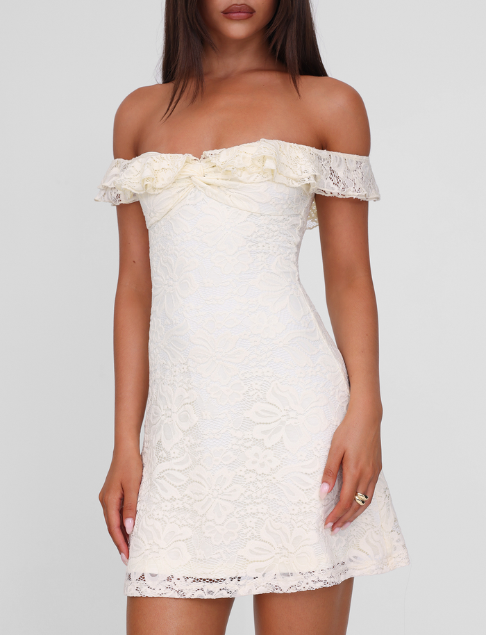 A woman wears the TIGERMIST DENALI LACE MINI DRESS in cream lace—an off shoulder mini dress with a ruffled neckline and fitted waist—standing against a plain light background.