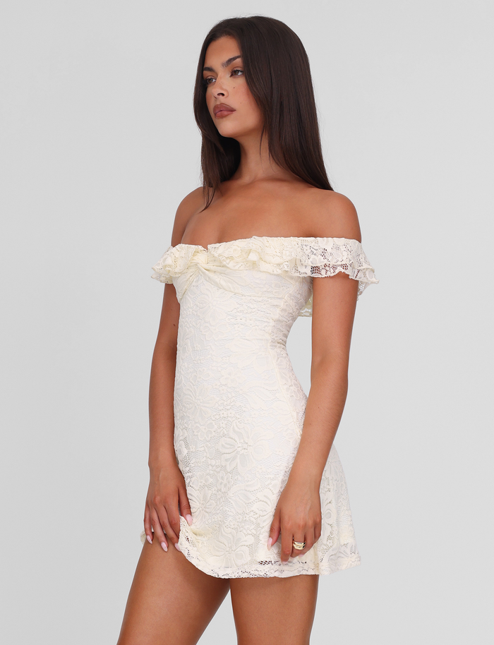 A woman with long, straight brown hair and a neutral expression stands against a plain background wearing the TIGERMIST DENALI LACE MINI DRESS in cream lace, featuring off-shoulder ruffled sleeves and delicate stretch lace.