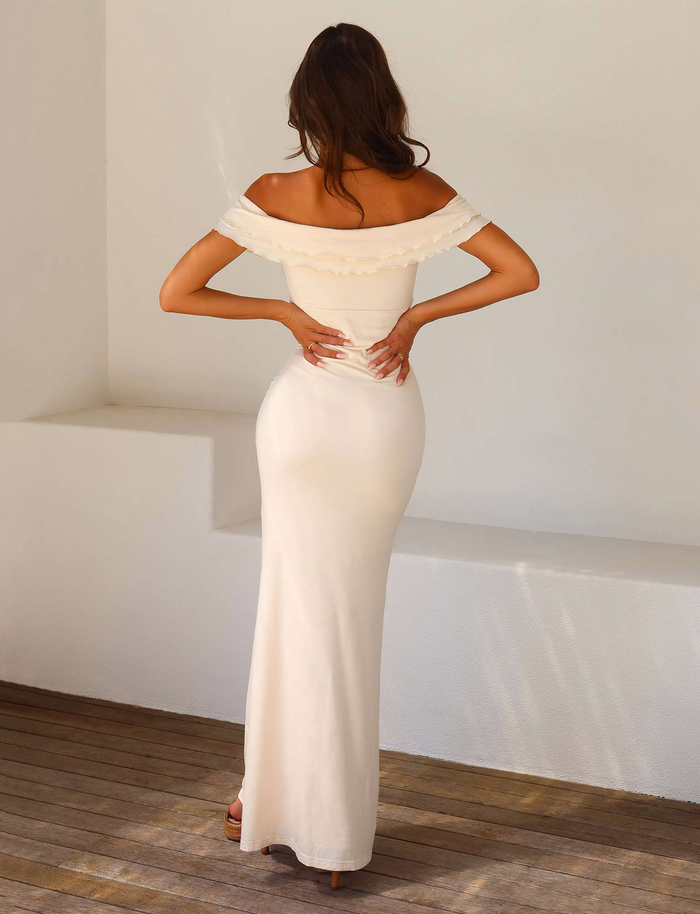 A woman with wavy brown hair stands indoors facing away, wearing the TIGERMIST DENALI MAXI DRESS - WHITE : BONE, an off-the-shoulder, sweetheart neckline bodycon maxi, paired with heeled sandals and hands resting on her lower back.