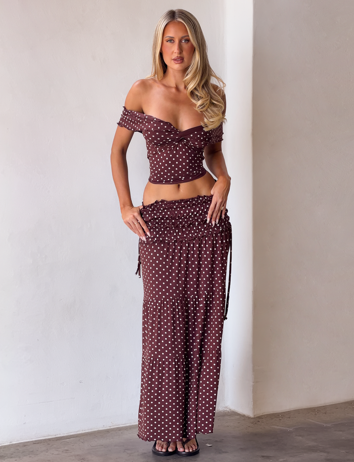 A woman with long blonde hair wears the TIGERMIST DENALI MAXI SKIRT - BROWN DOT and an off-shoulder crop top, both in soft modal fabric, indoors against a plain white wall.
