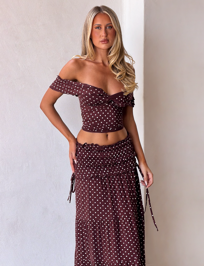 A woman with long blonde hair poses against a white wall, wearing the TIGERMIST DENALI MAXI SKIRT in brown dot print, featuring a shirred waistband and an off-shoulder brown crop top.
