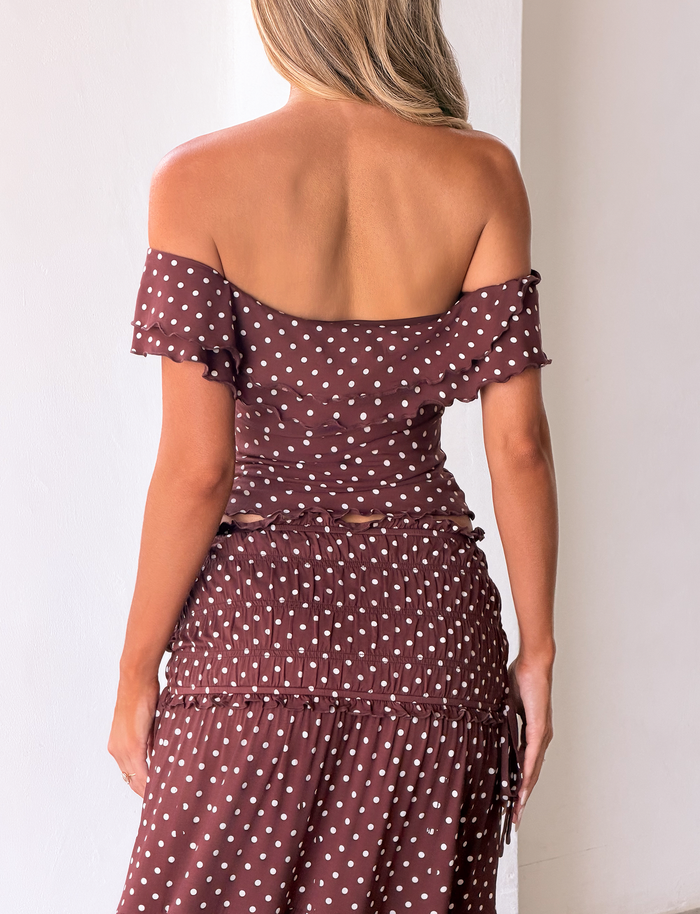 A woman stands with her back to the camera, wearing the TIGERMIST DENALI MAXI SKIRT - BROWN DOT and an off-the-shoulder top. Her loose, wavy light brown hair contrasts with the plain white background.