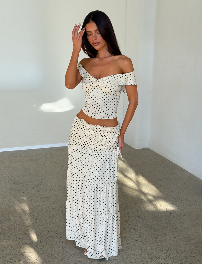 A woman stands in a sunlit room, wearing the TIGERMIST DENALI TOP - CREAM DOT and a matching long skirt, both cream with black polka dots. She touches her hair and looks down, casting a shadow on the speckled floor.