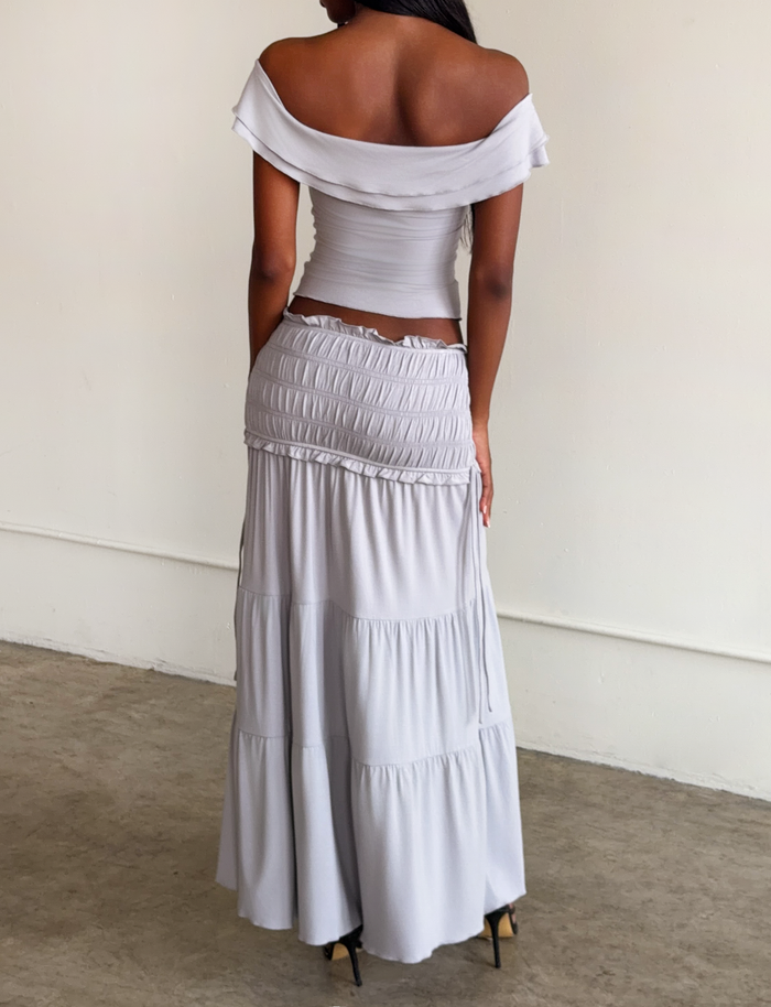 A woman with dark skin and straight hair stands indoors on concrete, wearing the TIGERMIST DENALI TOP in Dove Grey—an off-shoulder style with ruffled sleeves—paired with a matching long, tiered skirt.