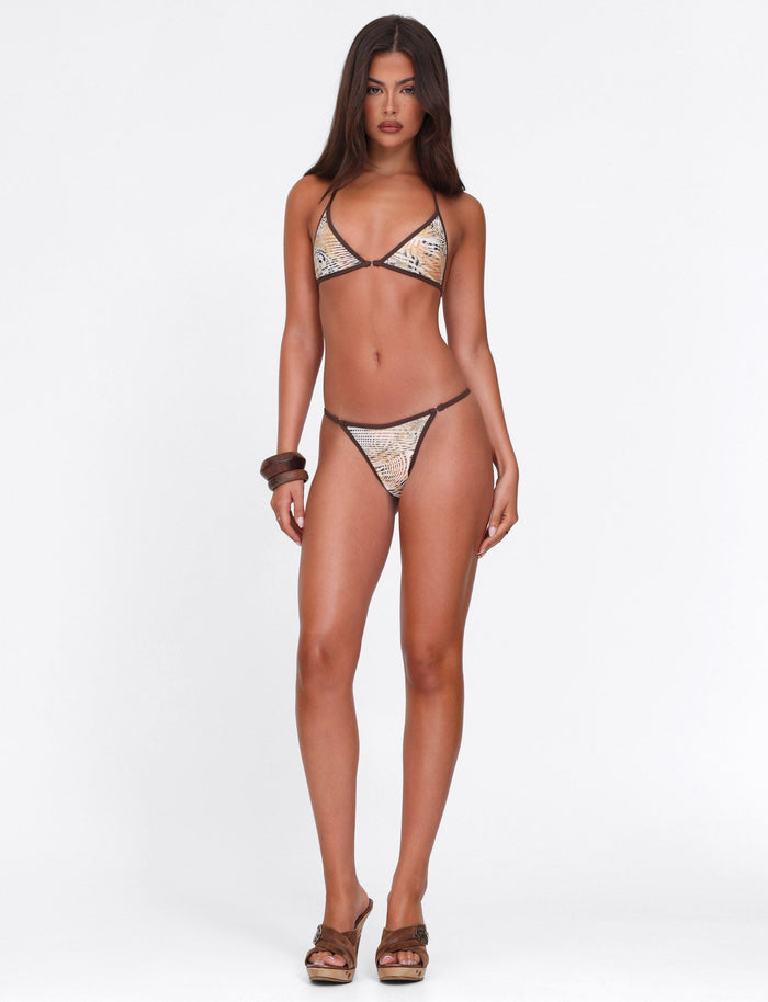 A woman with long brown hair stands against a white background, wearing the TIGERMIST DIANA BIKINI BOTTOM in Neutral Animal Marble, paired with brown sandals and a chunky bracelet. She faces forward with a neutral expression.