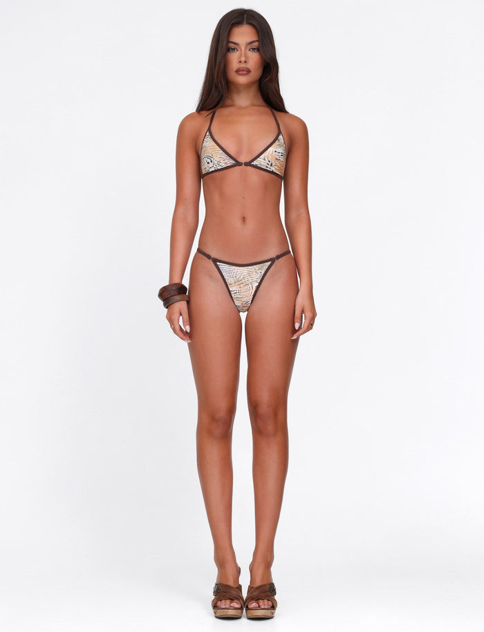 A woman with long brown hair stands facing forward in the TIGERMIST DIANA BIKINI TOP - NEUTRAL : ANIMAL : ANIMAL MARBLE, paired with brown heels and bracelets, against a plain white background. She has a neutral expression.