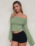 A woman with long, wavy blonde hair is looking to the side. She is dressed in the DIEDRE JUMPER - GREEN by TIGERMIST, an off-the-shoulder, long-sleeved, light green high stretch cotton top paired with high-waisted black shorts. The background is plain and light-colored.