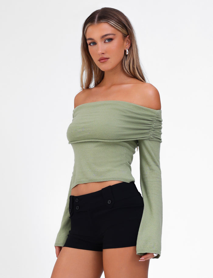 A woman with long, straight hair is wearing the TIGERMIST DIEDRE JUMPER - GREEN, an off-the-shoulder light green long-sleeve top made of high stretch cotton, paired with black high-waisted shorts. She is standing against a plain white background, looking slightly to her left with a neutral expression.