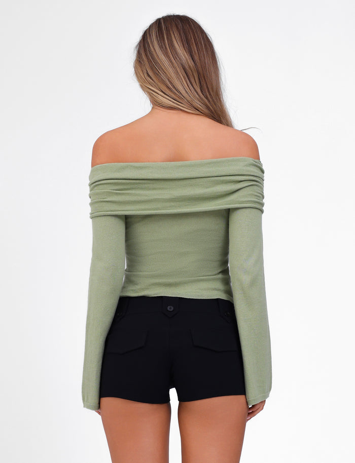 A woman with long hair stands with her back to the camera against a plain white background. She is wearing the DIEDRE JUMPER in green from TIGERMIST, an off-the-shoulder, long-sleeve fitted semi-sheer knit top paired with black high-waisted shorts.