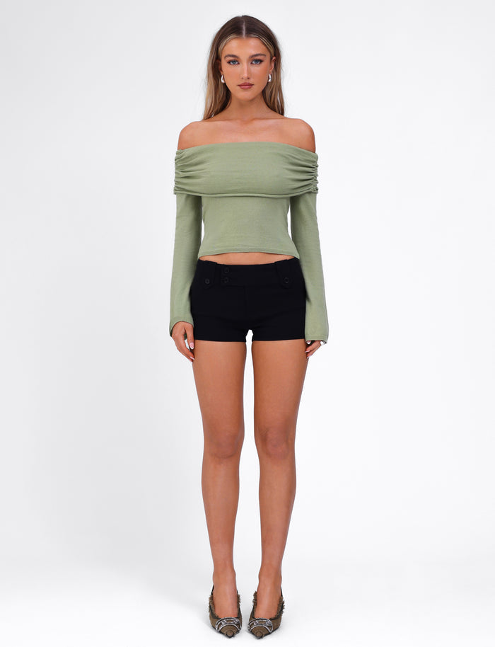 A woman stands against a plain background, wearing the TIGERMIST DIEDRE JUMPER in green with long sleeves and black high-waisted shorts made from high-stretch fabric. She has long, straight hair and is wearing patterned high heels. She is facing the camera directly with a neutral expression.