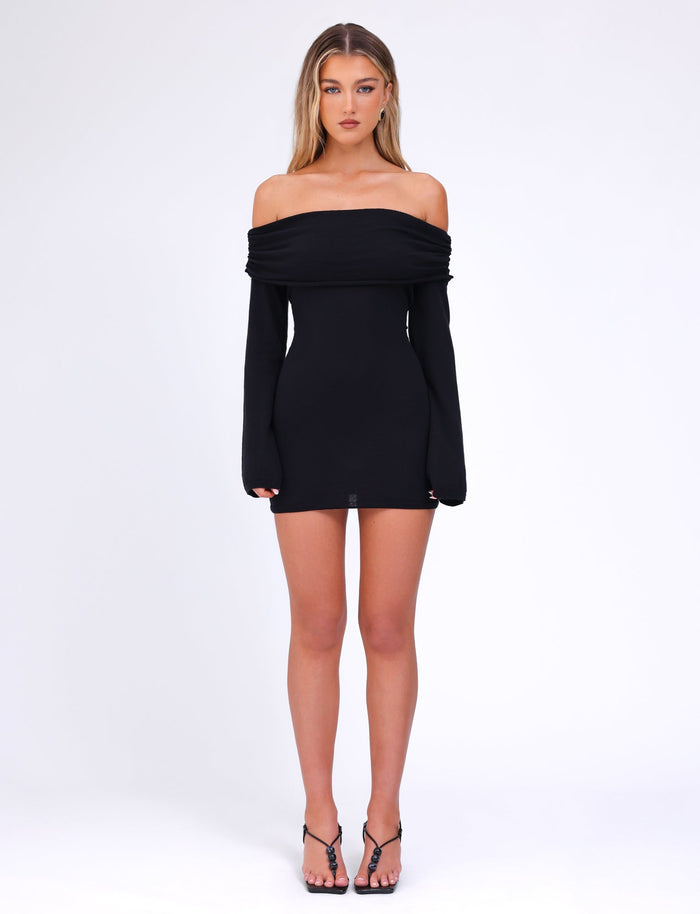 A woman stands against a white background, wearing an off-the-shoulder, long-sleeved Diedre Jumper Dress in black from TIGERMIST, paired with black strappy high-heeled sandals. She has long, straight hair and is looking directly at the camera.