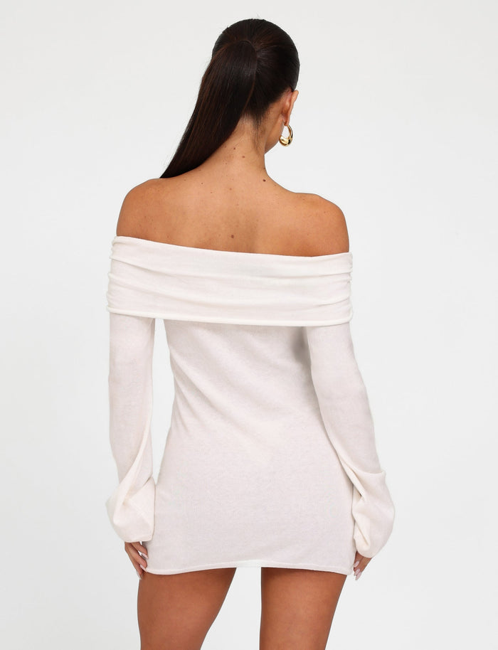 A woman with long, dark hair tied back is seen from behind, donning a sleek, off-the-shoulder Diedre Jumper Dress in white from TIGERMIST. The dress features long sleeves that extend past her wrists. She is also wearing gold hoop earrings, and the background is plain white.