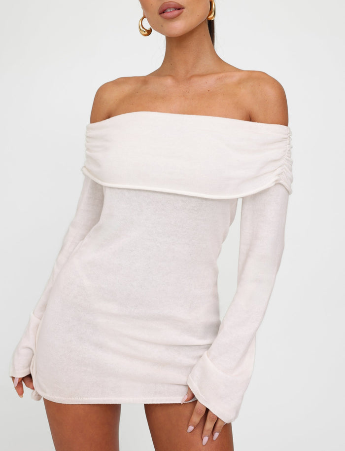 A woman is wearing the DIEDRE JUMPER DRESS - WHITE : CREAM from TIGERMIST. The off-the-shoulder dress features long sleeves, a fold-over collar, and extends to the mid-thigh. She is accessorizing with gold hoop earrings and standing against a plain white background.