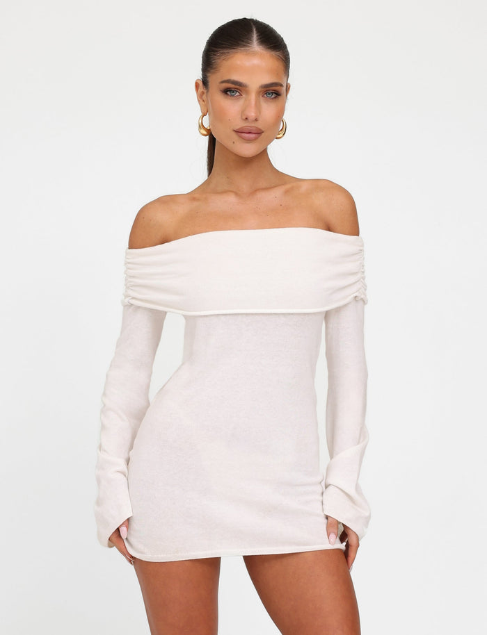 A woman stands against a white background, wearing the DIEDRE JUMPER DRESS - WHITE : CREAM from TIGERMIST. She has long, straight hair pulled back and is accessorized with large hoop earrings. Her hands rest relaxed on the dress.