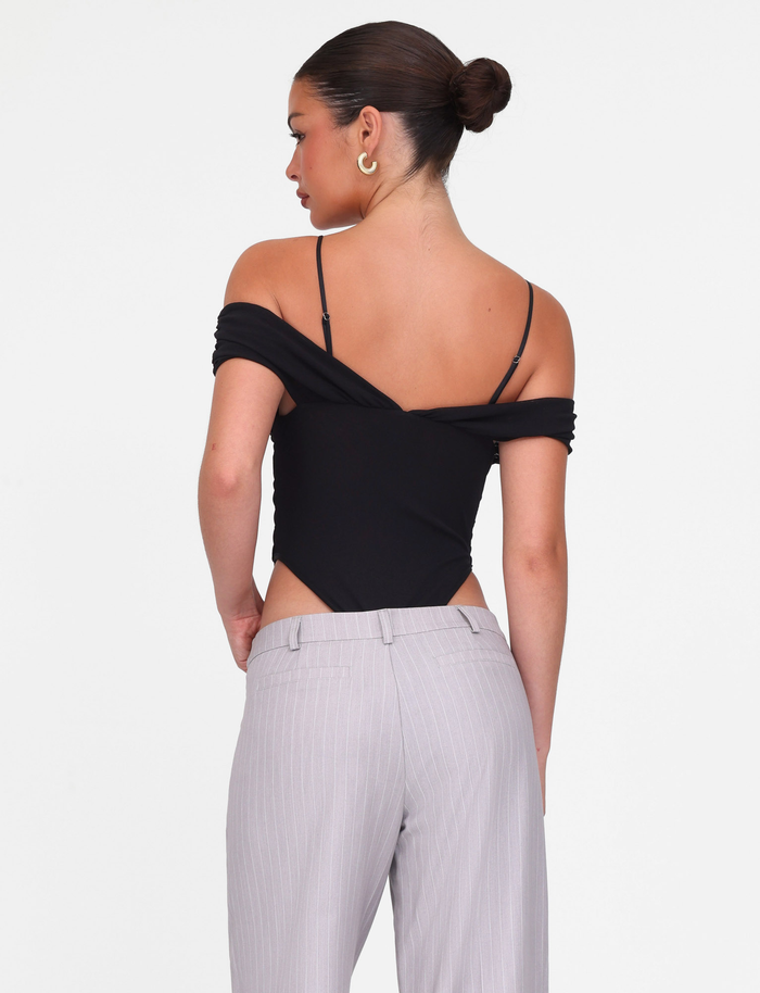A woman faces away, wearing the TIGERMIST DRAYA BODYSUIT in black, featuring off-the-shoulder ruched bust detail, paired with light grey pinstriped pants. Her dark hair is in a neat bun, and she wears large hoop earrings. White background.