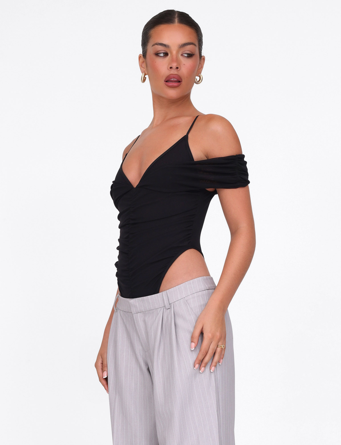 A woman models the TIGERMIST DRAYA BODYSUIT in black, featuring off-shoulder sleeves and thin straps, paired with light gray pinstriped pants against a plain white background, gazing slightly to the side.