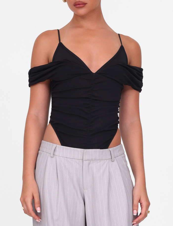 A woman wears the TIGERMIST DRAYA BODYSUIT in black with ruched bust and off-shoulder sleeves, paired with light gray pinstriped trousers, standing against a plain white background.