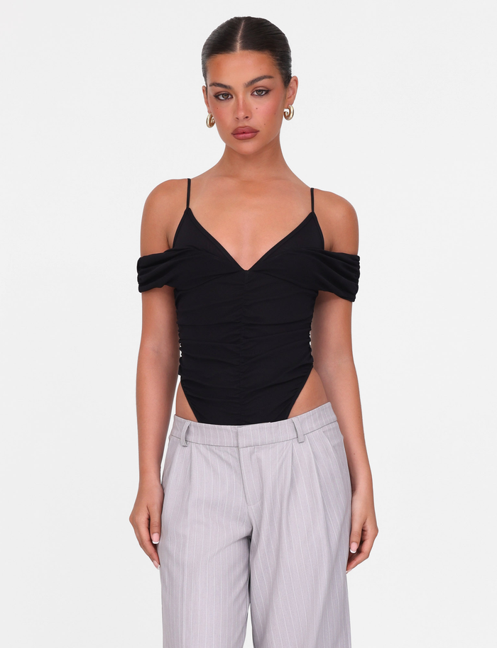 A woman with slicked-back hair models the TIGERMIST DRAYA BODYSUIT - BLACK, featuring off-shoulder sleeves and thin straps, paired with high-waisted light gray pinstripe trousers, against a plain white background.