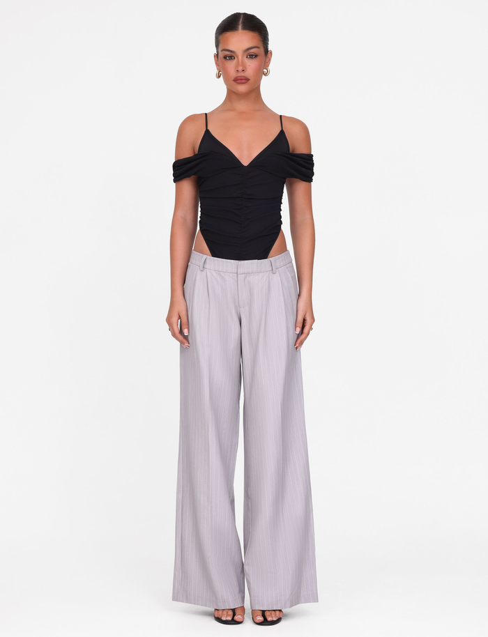 A woman faces forward against a white background wearing the TIGERMIST DRAYA BODYSUIT in black, featuring mesh fabric, ruched bust, and off-shoulder sleeves. She pairs it with high-waisted, wide-leg light gray trousers and black open-toe heels.