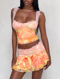 A woman stands against a plain light background, wearing the Tiger Mist USA CL - DUNE MESH MINI SKIRT in pastel orange and yellow with floral lace trim, paired with a matching camisole.