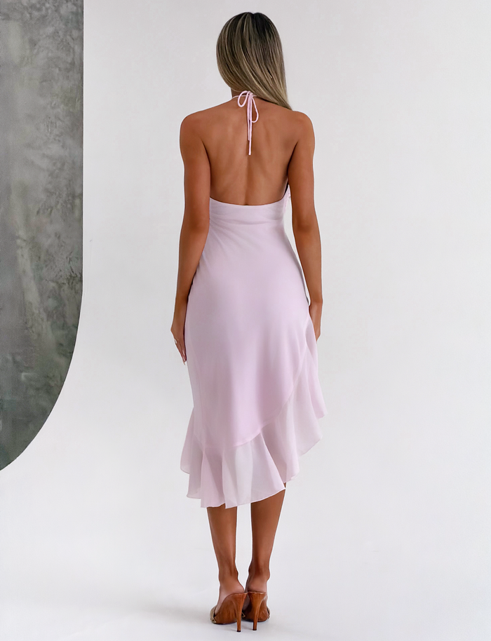 A woman faces away, modeling the TIGERMIST ELARINA MIDI DRESS in pink, featuring a halter-neck, backless design and asymmetrical hemline, paired with brown heeled sandals against a neutral background.