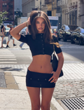A woman in a black crop top and mini skirt stands confidently on a city street, layered with the ELSIE CARDIGAN - BLACK by TIGERMIST. The vibrant city backdrop of cars, people, and buildings highlights her effortlessly chic style.