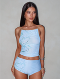 A woman with long, dark hair wears the ELVERA TOP - BLUE : GRADIENT FLORAL and matching shorts from TIGERMIST. She has neutral makeup and is accessorized with silver hoop earrings and silver bangles on her right wrist. She poses against a plain, light gray background.