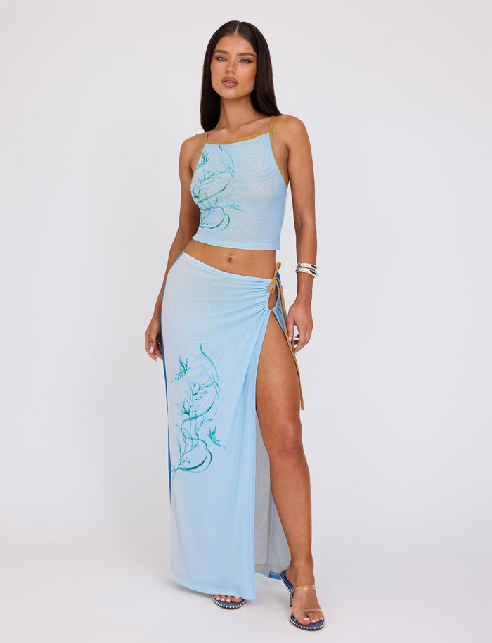 A woman is modeling the ELVERA TOP - BLUE : GRADIENT FLORAL from TIGERMIST, paired with a matching high-slit maxi skirt. The outfit features delicate floral designs. She stands with one hand on her hip, wearing silver bracelets and heeled sandals against a plain white background.