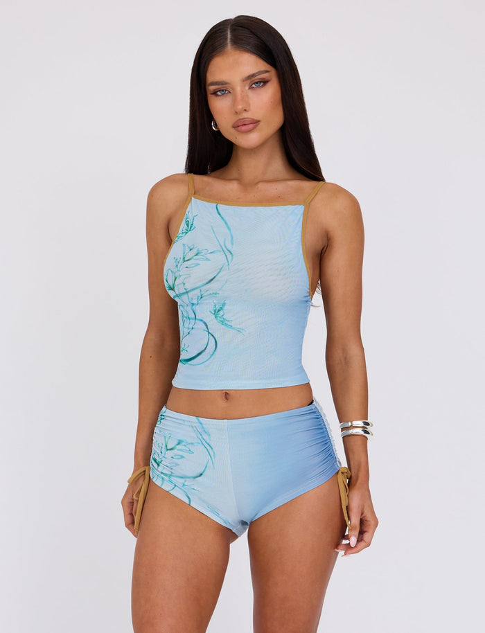 A person with long dark hair is wearing the ELVERA TOP - BLUE : GRADIENT FLORAL and matching blue shorts from TIGERMIST. They have their hands by their sides and are looking directly at the camera against a plain white background.