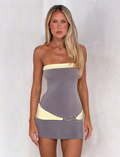 A woman with long blonde hair wears the TIGERMIST EOS MINI DRESS in grey and yellow, featuring a strapless, fitted silhouette and decorative hip belt, standing before a light textured background.