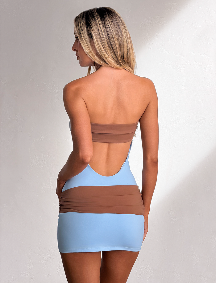 A woman with long blonde hair stands sideways against a white textured wall, wearing the TIGERMIST EOS MINI DRESS in soft blue and mocha featuring a strapless, color-blocked design with an open back and ruched detailing.