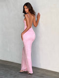 A woman in a form-fitting, backless ERICKA DRESS - PINK from TIGERMIST poses against a plain wall. She is facing sideways, with one hand touching the wall and looking at the camera. She is wearing heels and has long, wavy brown hair.