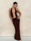 The woman poses elegantly against a textured beige wall, wearing a TIGERMIST ESHA MAXI SKIRT in chocolate brown with an asymmetrical waistband. Her hair is styled neatly, and she pairs the look with white sandals, one hand on her hip and the other gracefully on her leg.