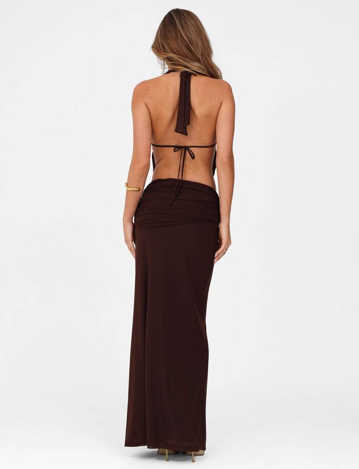 A woman with long hair, facing away, dons the elegant TIGERMIST Esha Maxi Skirt in chocolate—a dark brown backless halter-neck style tied gracefully at the back. Complemented by a gold bracelet on her left arm and heels, her poised silhouette is accentuated against a plain white background.