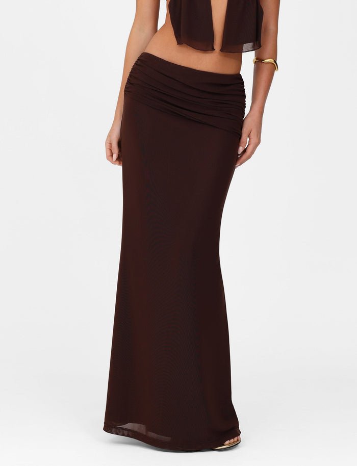 A person in a TIGERMIST ESHA Maxi Skirt in chocolate brown with a ruched A-line, asymmetrical waistband, paired with a sleeveless black top that reveals some midriff, accessorized by a gold cuff bracelet on their right wrist.