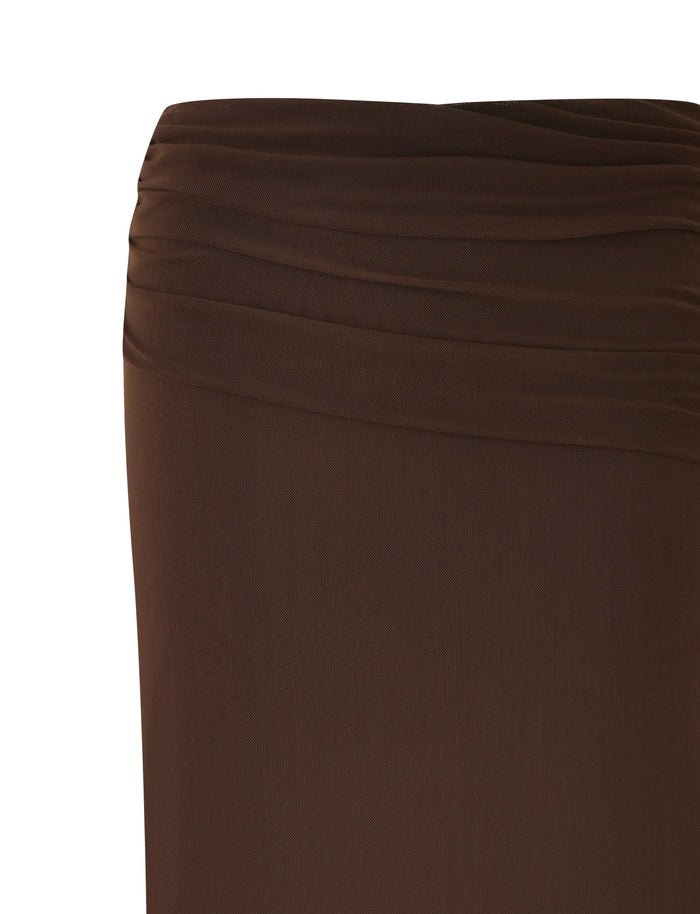 A close-up of the ESHA MAXI SKIRT - BROWN : CHOCOLATE by TIGERMIST, showcasing its knee-length style with an asymmetrical waistband that enhances its draped design and elegant pleat details. The fabric appears smooth and lightweight, exuding sophistication.