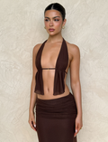 A woman with dark hair in a sleek bun wears the TIGERMIST ESHA TOP - BROWN : CHOCOLATE with adjustable ties and a matching skirt, standing against a light textured wall and looking calmly to the side.