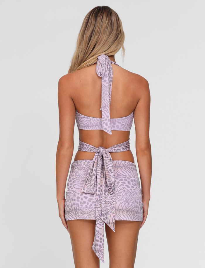 A woman with long blonde hair is seen from the back wearing the TIGERMIST EVANGELINE TOP in Pale Pink Tiger print and a matching mini skirt with wrap tie details, both standing out against a plain white background.