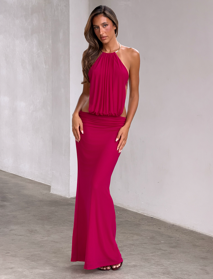 A woman poses indoors in the TIGERMIST EVANNA MAXI DRESS - BERRY, a sleeveless, floor-length mesh gown with a gold trim halter neckline. She stands gracefully with long, wavy hair and her hands at her sides.