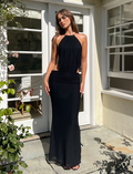 A woman stands on a stone patio in the TIGERMIST EVANNA MAXI DRESS - BLACK, posing before white French doors with sunlight and plants around her, a room visible inside the house.