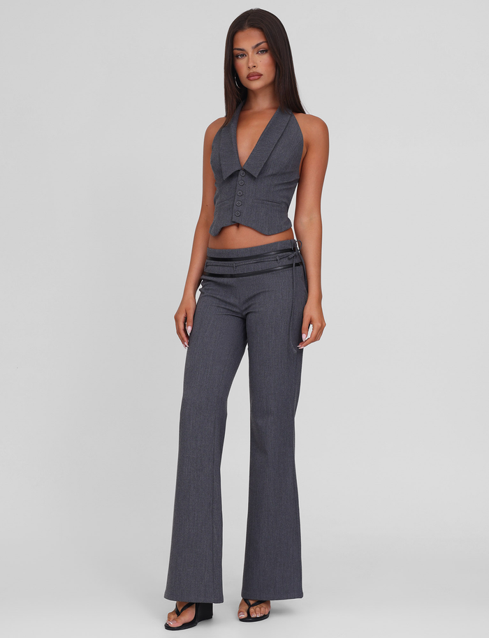 A woman with straight dark hair and a neutral expression wears TIGERMIST's FEDRA PANT - GREY, styled with a sleeveless deep V-neck grey vest featuring PU leather trim and black heeled sandals, against a plain light background.