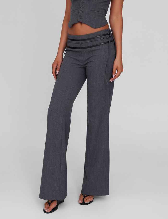 Wearing TIGERMIST’s FEDRA PANT in grey with PU leather trim and a matching cropped vest, a person in black sandals poses against a plain light gray background. Only their lower torso, legs, and one arm are visible.