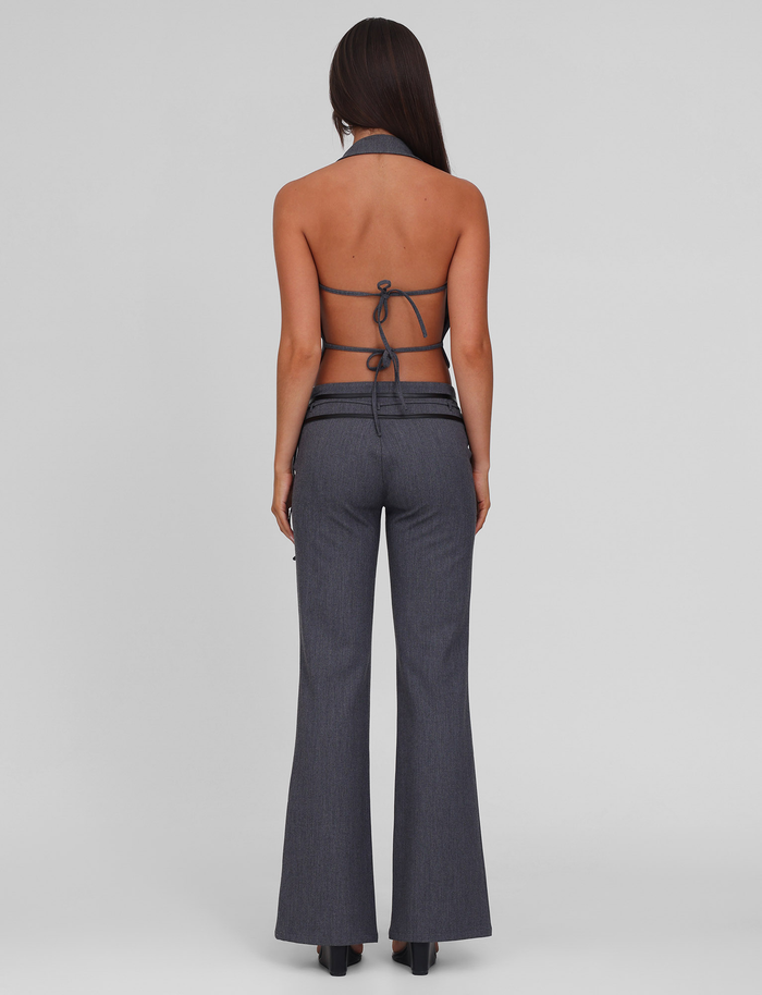 A woman with long straight hair faces away, modeling the TIGERMIST FEDRA PANT in grey and a matching halter-neck top with PU leather trim, tied at the back to reveal her back, against a plain light grey background.