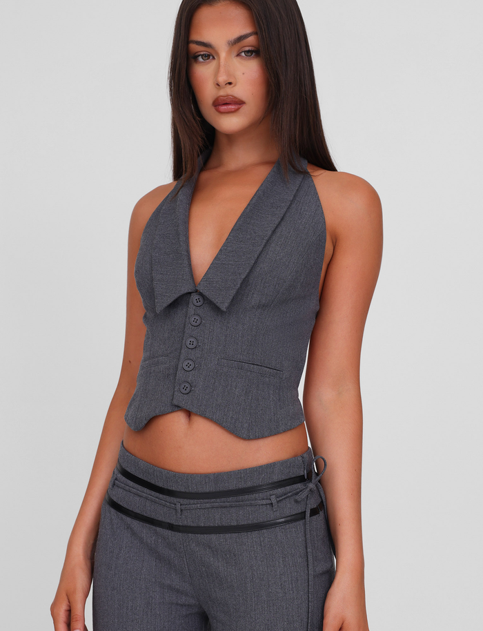 A woman with long brown hair models the TIGERMIST FEDRA VEST - GREY, a stylish backless halter vest with buttons, paired with matching grey pants against a plain light background.