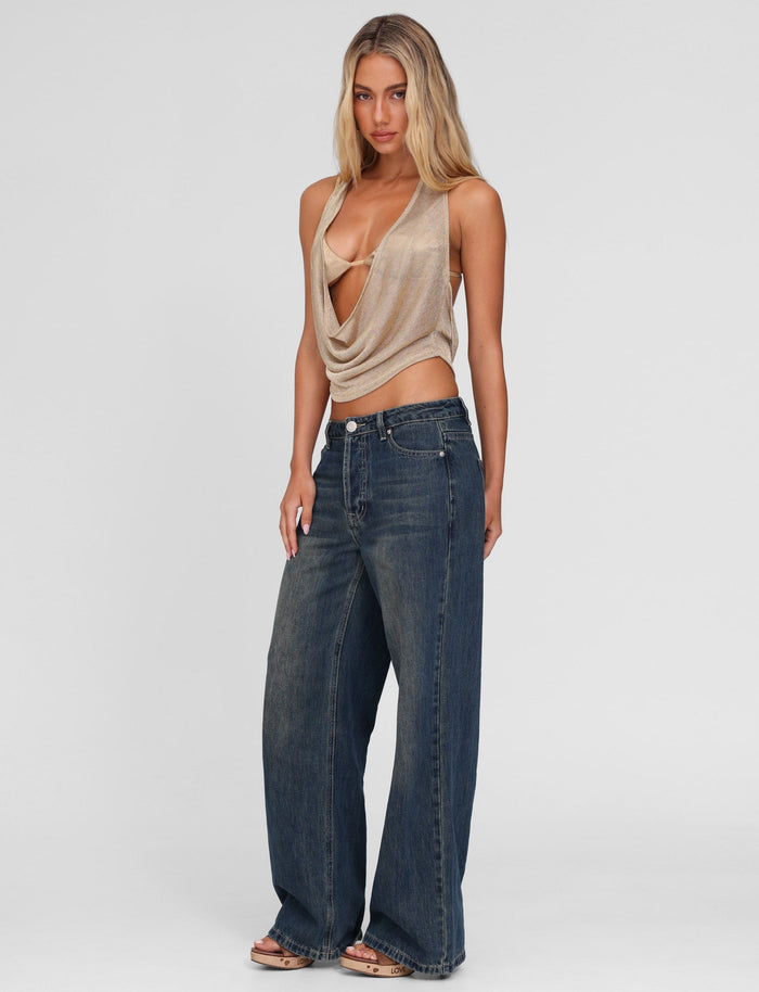 A woman with long, wavy blonde hair stands against a plain background wearing TIGERMIST's FELIX JEAN - BLUE: MID WASH, styled high-waisted and wide-leg, paired with a beige draped halter top. She looks directly at the camera with a neutral expression.