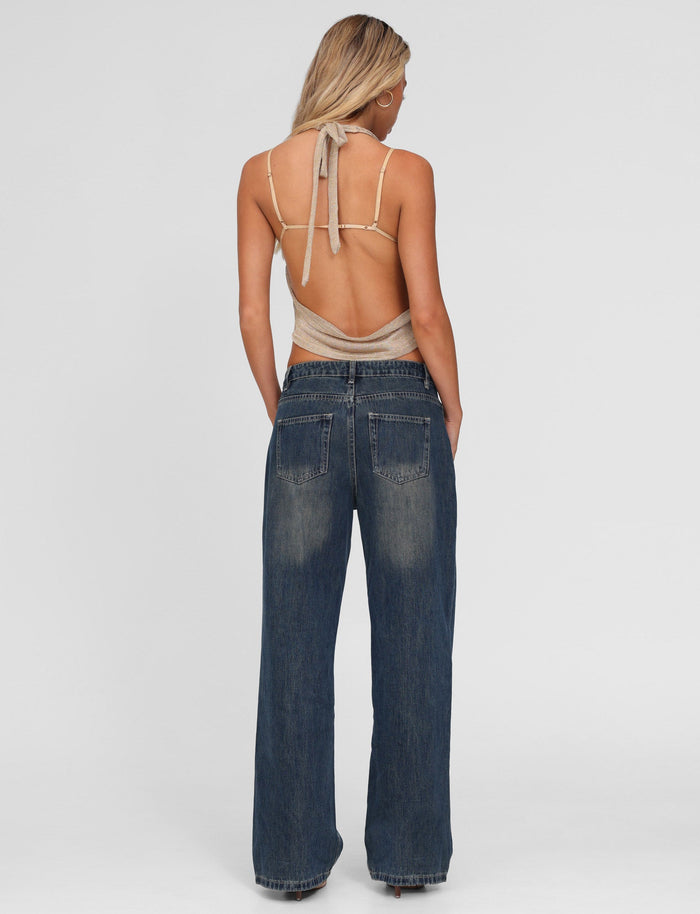 A woman with blonde hair stands facing away, wearing TIGERMIST's FELIX JEAN - BLUE : MID WASH and a beige halter top with an open back. Her hands rest in her pockets as her hair falls over her shoulders.