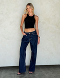 A woman with long blonde hair poses against a light textured wall, wearing a black sleeveless crop top and TIGERMIST FELIX JEAN - DARK DENIM wide leg pants, with one hand in her pocket and the other by her side.