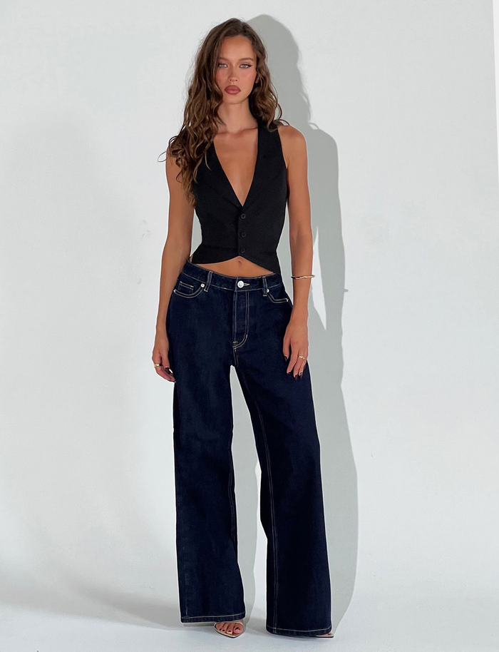 A woman with long wavy hair poses confidently against a white background, wearing TIGERMIST's FELIX JEAN - DARK DENIM in an oversized, high-waisted wide-leg fit and a sleeveless black vest.