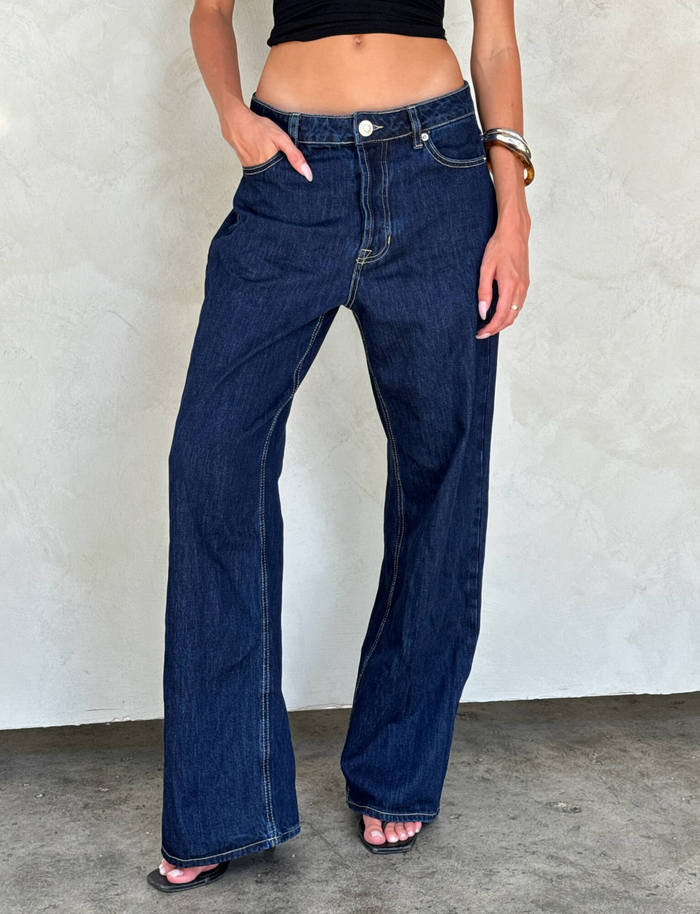 A person wears TIGERMIST's FELIX JEAN - DARK DENIM wide-leg jeans, paired with a black crop top and open-toe heels, posing against a light-textured wall with one hand in their pocket.