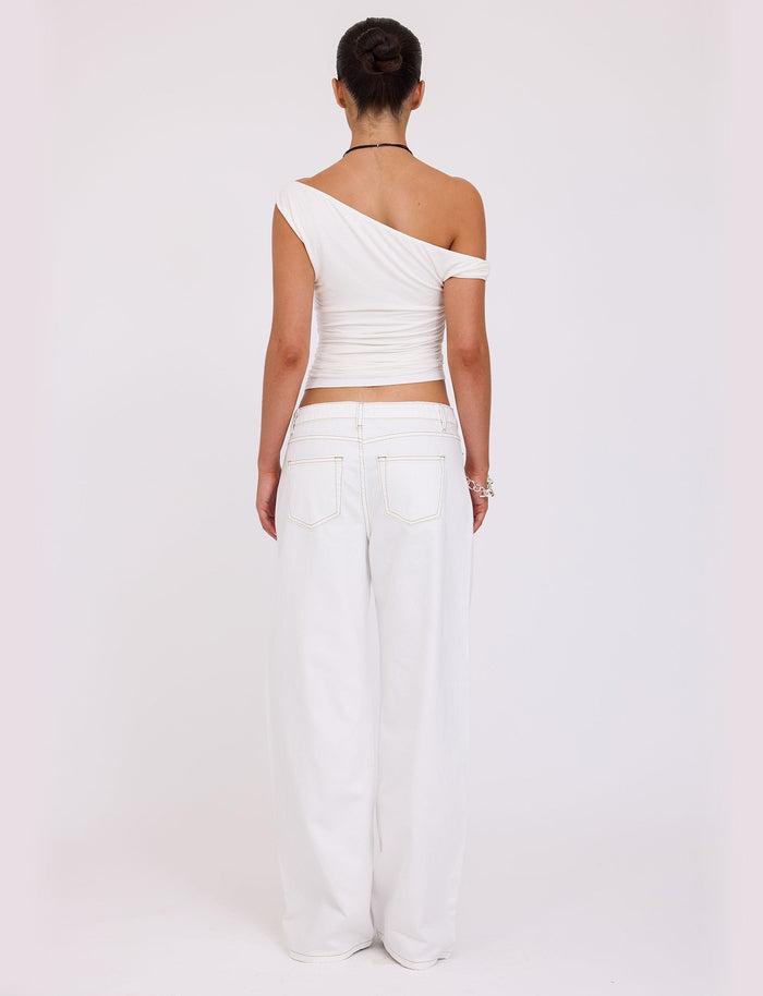 A person is standing with their back to the camera, stylishly dressed in the Tiger Mist FELIX JEAN - WHITE, featuring a one-shoulder top and high-waisted, wide-leg pants. Their hair is styled in a neat bun, complemented by a bracelet and necklace.